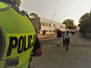 Policjant stoi tyłem w odblaskowej kamizelce. W tle dzieci idące na rozpoczęcie roku szkolnego.