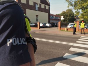 Bok koszuli służbowej policjanta. W tle, przed przejściem dla pieszych stoi policjant wraz z funkcjonariuszem Żandarmerii Wojskowej.