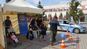 Policjanci podczas prezentacji stoiska policyjnego. Obok stoiska stoi radiowóz. Odwiedzający stoisko korzysta z toru przeszkód używając alkogogli.