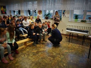 Policjantka kuca przy chłopcu, który tak jak reszta uczniów siedzi na ławce i słucha pogadanki o tematyce bezpiecznych zachowań zimowych.