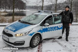 Starszy sierżant Michał Niziałek ubrany w czarny policyjny mundur stoi przy otwartych drzwiach radiowozu na tle jeziora i ośnieżonego lasu.