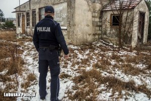 Policjant w granatowym mundurze stoi tyłem do zdjęcia na tle ośnieżonych pustostanów.