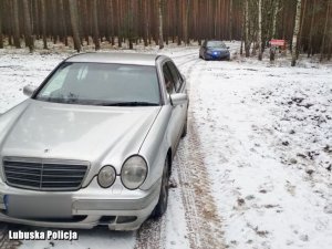 Srebrny mercedes stoi na ośnieżonej leśnej drodze. W tle widoczny jest nieoznakowany radiowóz.