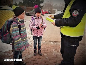 Policjant w odblaskowej kamizelce wręcza dziewczynkom odblaskowe opaski. Sytuacja ma miejsce na ośnieżonej drodze tuż przed szkołą.