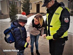 Policjant w odblaskowej kamizelce wręcza chłopcu odblaskową opaskę. Nad obdarowanym nachyla się jego mama. Sytuacja ma miejsce na ośnieżonej drodze tuż przed szkołą.