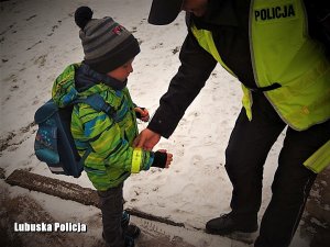 Policjant w odblaskowej kamizelce zakłada chłopcu w ubiorze zimowym odblaskową opaskę na rękę. Sytuacja ma miejsce na ośnieżonej drodze tuż przed szkołą.