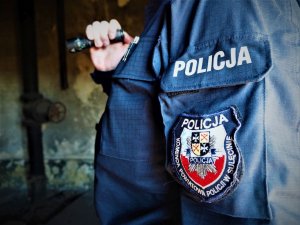 Policjant w pustostanie trzyma w ręku latarkę. Widoczny jest bok jego mundury z naszywką identyfikującą jednostkę.