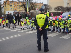 Policjant w kamizelce odblaskowej stoi tyłem do zdjęcia a przodem w kierunku przejścia dla pieszych, po którym poruszają się przedszkolaki również w odblaskowych kamizelkach.