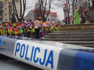 Celka policyjnego radiowozu. W tle przedszkolaki w odblaskowych kamizelkach z kolorowymi bibułowymi kwiatkami w dłoniach.