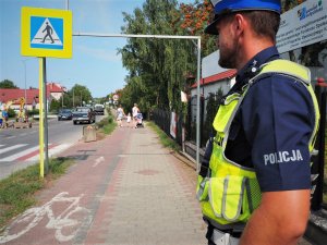 Policjant na tle przejścia dla pieszych oraz ścieżki rowerowej z rejonie szkoły.