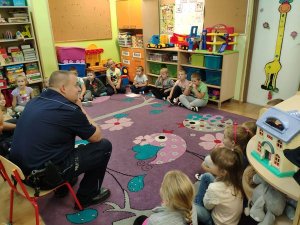 Policjant siedzi wraz z przedszkolakami w kole. Znajdują się w sali placówki.