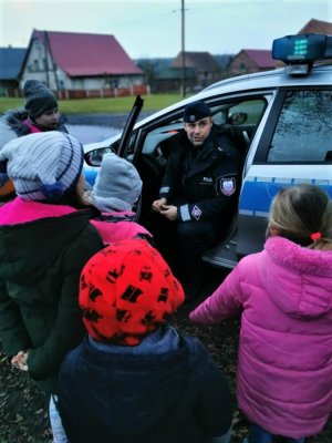 Dzielnicowy siedzi w aucie, dzieci stoją przed radiowozem. Mundurowy wyjaśnia jak uruchomić światła błyskowe.