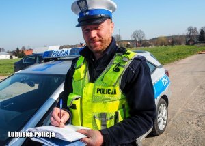 Policjant ruchu drogowego piszący w notatniku służbowym. W tle radiowóz.