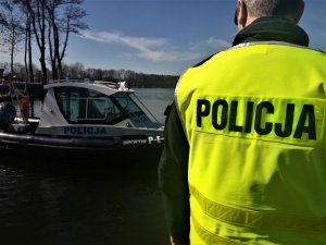 Policjant stojący tyłem w kamizelce odblaskowej. Na drugim planie łódź policyjna.