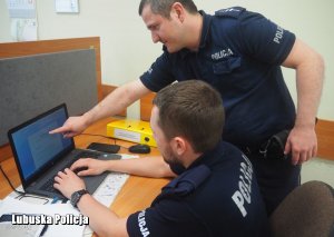 Dzielnicowy tłumaczy młodemu policjantowi siedzącemu przy komputerze, jak sporządzać pisma urzędowe.