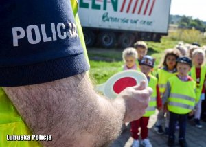 Na pierwszym planie znajduje się dzielnicowy stojący bokiem. Trzyma w rękach tarczę do zatrzymywania pojazdów. Na drugim tle znajdują się przedszkolaki w kamizelkach odblaskowych.