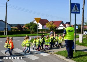 Dzieci pod czujnym okiem dzielnicowego przechodzą w parach przez przejście dla pieszych.