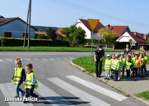 Dzieci przechodzą w parach przez przejście dla pieszych. Wszystkiemu przygląda się dzielnicowy.