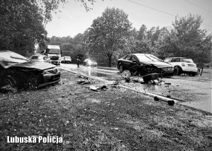 Czarno białe zdjęcie przedstawiające drogę krajową numer 22, na której znajdują się znacznie uszkodzone pojazdy biorące udział w zdarzeniu drogowym.