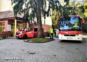 Przed budynkiem szpitala stoi autobus i bus straży pożarnej. Przy nim stoją medycy ubrani w kombinezony oraz żołnierze Wojsk Obrony Terytorialnej.