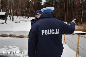 Policjanci stoją na pomoście. Jeden z nich bokiem, zaś drugi stoi tyłem do zdjęcia wskazując palcem na jezioro.