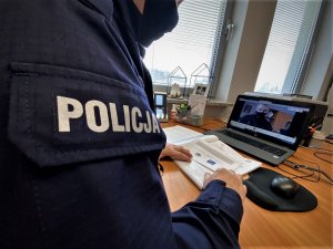 Bok rękawa policyjnego mundury z napisem Policja. Na dalszym planie biurko i stojący na nim laptop. Policjant trzyma w ręku dokumentację.