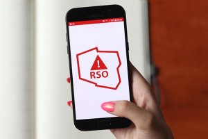 W kobiecej dłoni znajduje się telefon komórkowy. Na jego ekranie, na białym tle wyświetlany jest czerwony kontur Polski, wewnątrz którego znajduje się znak wykrzyknika wpisany w trójkąt, a pod nim napis RSO.
