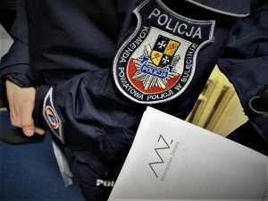 Na pierwszym planie biała karta z logo Ministerstwa Zdrowia. Na drugim planie rękaw policyjnego kurtki z naszywką Komendy Powiatowej Policji w Sulęcinie.