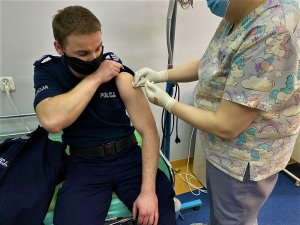 Policjant w maseczce odsłania ramię do przyjęcia szczepionki. Pani pielęgniarki wykonuje zastrzyk.