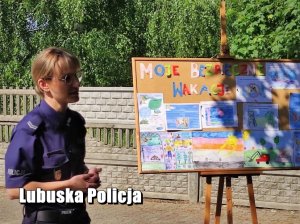 Policjantka podczas prowadzenia zajęć edukacyjnych dla dzieci na świeżym powietrzu. Obok stoi tablica z napisem Moje Bezpieczne Wakacje oraz rysunkami dzieci.