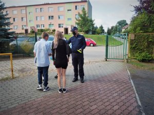 Policjant w maseczce ochronnej wręcza chłopcu i dziewczynce odblaskowe opaski na terenie szkoły.