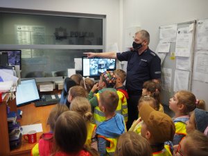 dzieci stoją w pomieszczeniu dyżurnego policji a policjant ręką wskazuje im na monitor komputera