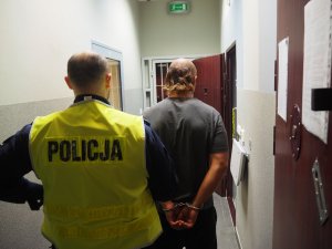 Policjant i mężczyzna stoją tyłem. Policjant trzyma mężczyznę za rękę a mężczyzna ma założone kajdanki