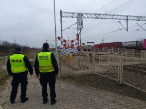 Policjant i funkcjonariusz Służby Ochrony Kolei idą chodnikiem obok torowiska