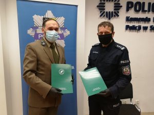 Umundurowany policjant stoi wraz z pracownikiem lasów państwowych na tle baneru sulęcińskiej policji i trzymają zielone aktówki