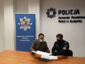 Umundurowany policjant siedząc wraz z pracownikiem lasów państwowych wymieniają się dokumentami