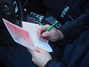 Policjant siedzi w radiowozie, sprawdza dokumenty autokaru. Robi zapisy w notatniku służbowym.