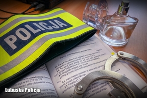 Na otwartym Kodeksie Karnym leży odblaskowa opaska naramienna z napisem Policja, poniżej kajdanki. Po prawej stronie kodeksu znajduje się flakon perfum.