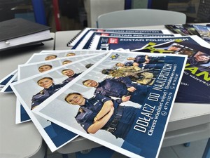 Na stole leżą ulotki promujące służbę w Policji z hasłem &quot;Dołącz do najlepszych&quot;.