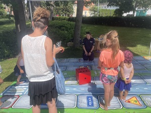 Dzieci grają w wielkoformatową grę edukacyjną. Policjanta klęka przy planszy, nadzorując przebieg rozgrywek.