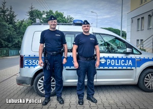 Od lewej sierżant sztabowy Piotr Sławiak oraz posterunkowy Michał Banaszak stoją przed radiowozem. Ubrano się w policyjne mundury oraz furażerki.