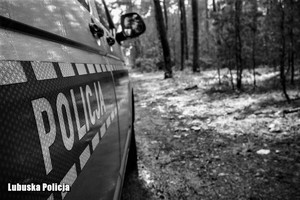 Bok radiowozu z napisem Policja na tle terenu leśnego. Zdjęcie jest czarno-białe.