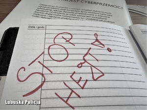 W policyjnym notatniku widnieje czerwony napis STOP HEJT! W tle książka z definicją cyberprzemocy.