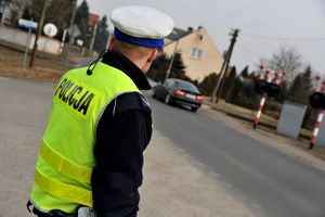 Policjant obserwuje ruch kołowy w rejonie przejazdu kolejowego. Ma założoną odblaskową kamizelkę.