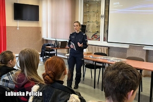 Policjantka prowadzi spotkanie promujące służbę w Policji z maturzystami. Uczniowie siedzą tyłem do zdjęcia w świetlicy szkolnej.