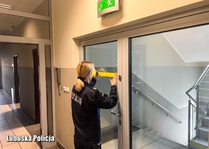 Policjantka nakleja żółtą taśmę na szklane skrzydło drzwiowe.