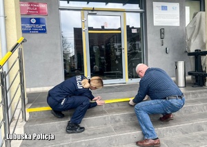 Funkcjonariuszka wraz z pracownikiem cywilnym oklejają koniec ciągu schodów zewnętrznych prowadzących do jednostki Policji.