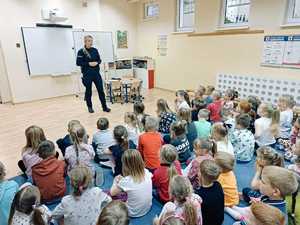 Policjantka prowadzi spotkanie edukacyjne dla grupy przedszkolaków. Dzieci siedzą po turecku w sali przedszkolnej, tyłem do zdjęcia.