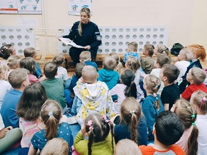 Policjantka prowadzi spotkanie edukacyjne dla grupy przedszkolaków. W rękach trzyma książkę. Dzieci siedzą po turecku w sali przedszkolnej, tyłem do zdjęcia.