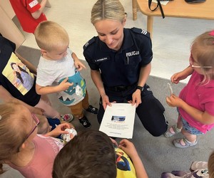 Policjantka kuca przy przedszkolakach, którzy wręczają jej podziękowanie za przeprowadzone spotkanie.